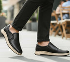 Person wearing black slip-on shoes with beige soles on a blurred outdoor background