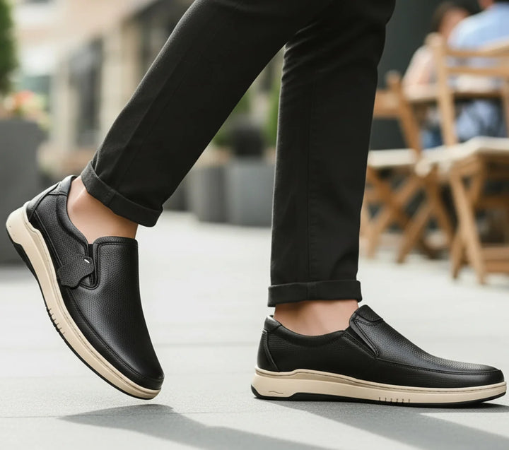 Person wearing black slip-on shoes with beige soles on a blurred outdoor background