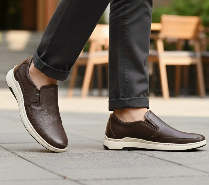 Brown slip-on shoes worn with black pants on a blurred outdoor background